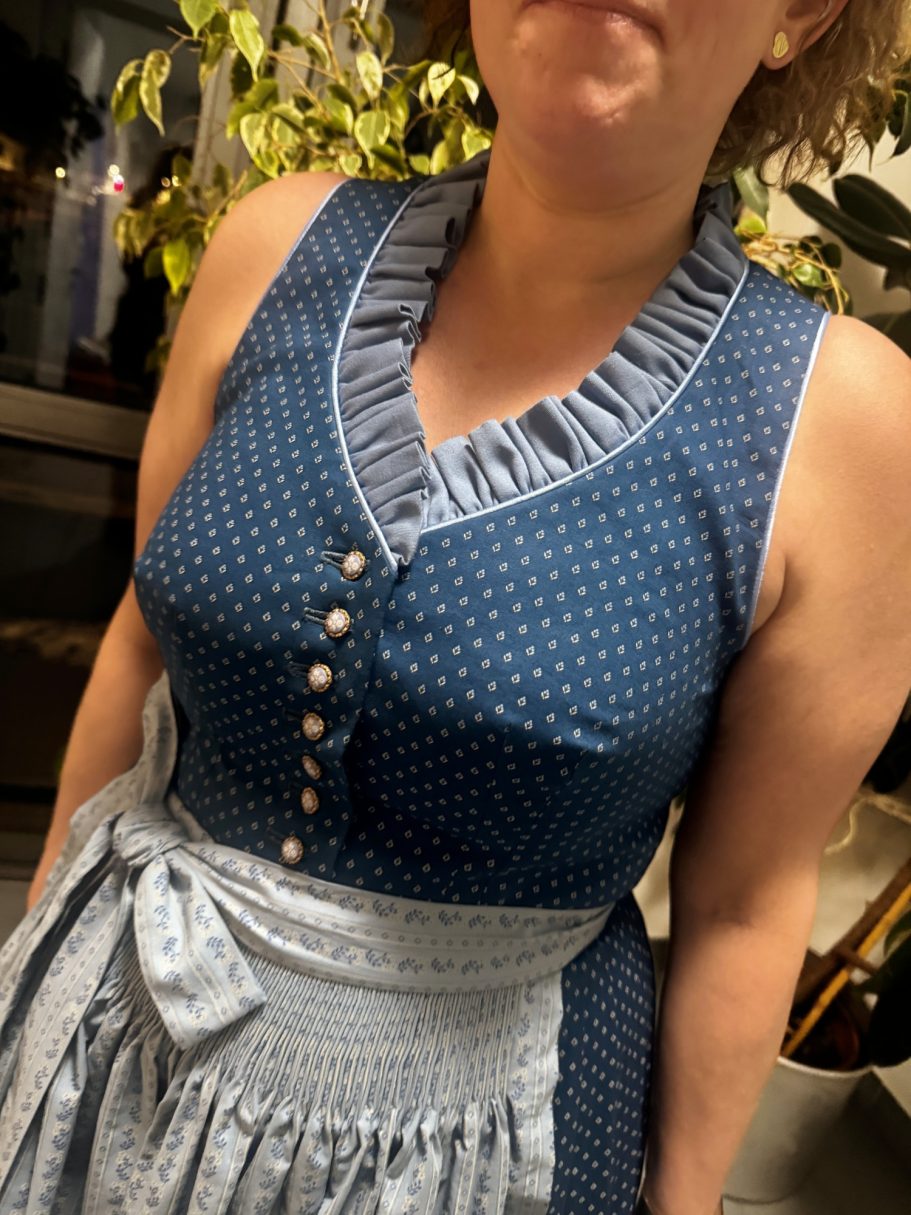 Frau in einem blauen dirndlförmigen Kleid mit Rüschen und Knöpfen vorne.